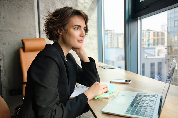Retrato de una mujer trabajadora en una oficina, usando un portátil y un teléfono móvil, sentada cerca de la ventana en un espacio de coworking. - Foto, Imagen