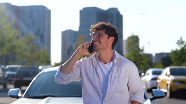 Улыбающийся парень в очках стоит возле своей машины, разговаривая по мобильному телефону в городе. Транспорт, технологии и образ жизни. Медленное движение - Кадры, видео