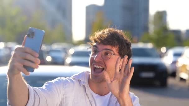 Un gars souriant qui utilise son téléphone portable, fait du selfie, agite la main et montre un signe en V. Voiture en arrière-plan. Transport, technologie et mode de vie concept. Mouvement lent - Séquence, vidéo