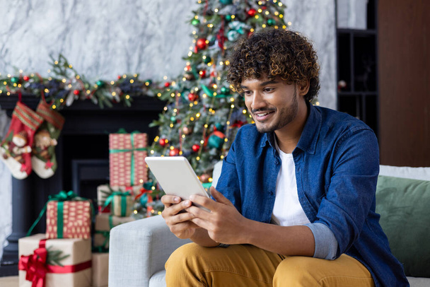 Een man gebruikt een tablet computer terwijl hij thuis zit in de woonkamer met Kerstmis. Een Latijns-Amerikaan gebruikt de app, bekijkt een video, glimlacht, viert nieuwjaarsvakantie in de buurt van de kerstboom. - Foto, afbeelding