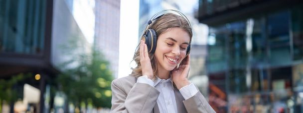 Πορτρέτο της χαμογελαστής εταιρικής γυναίκας, κορίτσι με κοστούμι ακούγοντας μουσική στα ακουστικά, κάθεται στο κέντρο της πόλης. - Φωτογραφία, εικόνα