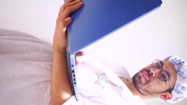 Video verticale di uomo che indossa asciugamano e cuffia da bagno guardando computer portatile blu  - Filmati, video