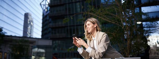 Mujer de negocios sonriente usando su teléfono móvil en un descanso, fuera de la oficina, mirando la pantalla del teléfono inteligente mientras envía mensajes a alguien. - Foto, imagen