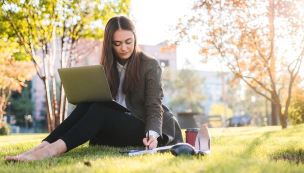 Dizüstü bilgisayarı olan esmer bir kadın güneşli bir parkta çimlerin üzerinde oturuyor ve not alıyor. Çevrimiçi eğitim, uzaktan çalışma ve iletişim - Fotoğraf, Görsel