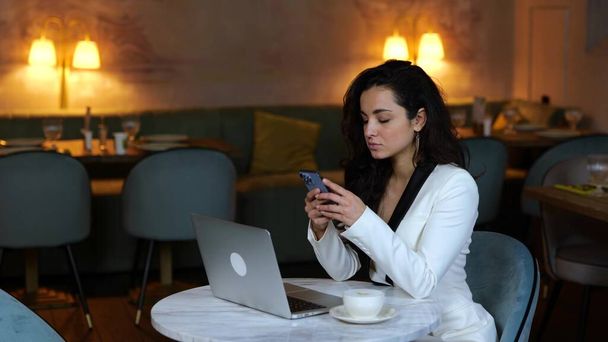 Elegancka młoda bizneswoman siedzi przy stole w restauracji podczas korzystania ze smartfona. Laptop i kawa na stole. Koncepcja biznesu. Zwolniony ruch - Zdjęcie, obraz