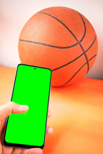 Mão segurando smartphone com tela verde ao lado de bola de basquete laranja - Foto, Imagem