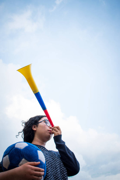 Mujer sosteniendo pelota de fútbol azul y utilizando vuvuzela para apoyar al equipo en un día nublado  - Foto, imagen