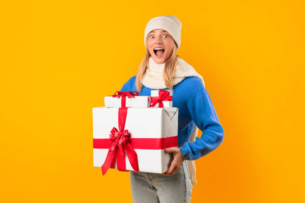 Захоплена молода жінка щасливо тримає стек різдвяних подарункових коробок, позуючи над яскравим жовтим студійним фоном, символізуючи щедрий дух свята і живі моменти святкування - Фото, зображення