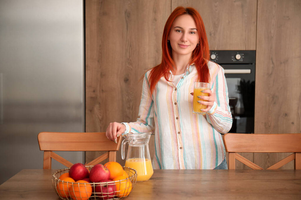 Молодая рыжая женщина с кувшином и стаканом сока на кухне - Фото, изображение