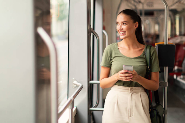 Pasażerka przeglądająca się przez telefon w tramwaju wewnątrz, stojąca ciesząc się podróżą po mieście w nowoczesnym tramwaju, patrząc na bok podczas pisania SMS-ów. Dojazd i łączność - Zdjęcie, obraz