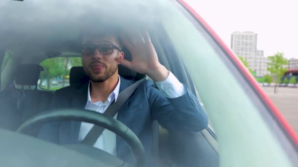 Joven feliz en gafas conduciendo un coche por la ciudad y viendo a un amigo, agitando la mano. Concepto de transporte, negocios y personas. Movimiento lento - Metraje, vídeo