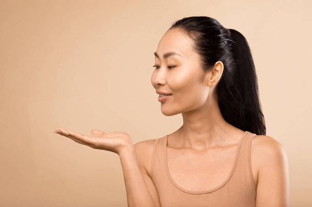 Femme asiatique d'âge moyen sereine, étendant la main et présentant un nouveau produit de beauté sur sa paume, tournée dans un studio avec une toile de fond beige doux - Photo, image
