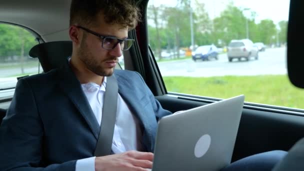 Nuori liikemies virallisissa vaatteissa kannettavan tietokoneen ja kirjoittamalla istuessaan takapenkillä autossa. Liiketoiminnan, matkan, liikenteen ja teknologian käsite. Hidastus - Materiaali, video