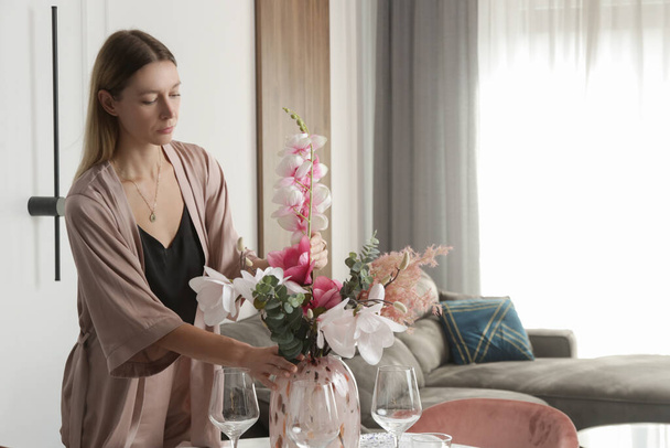 Woman arranging vase with flowers on dining table. Housewife taking care of coziness in apartment. Interior decor, household and home improvement concept.  - Photo, Image