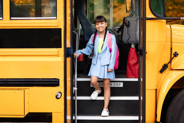 Lächelndes asiatisches Mädchen beim Aussteigen aus dem gelben Schulbus, fröhliches weibliches Kind mit Rucksack steigt aus dem Fahrzeug, als es zum Lernen ankommt, bereit für Unterricht und Lernspaß - Foto, Bild