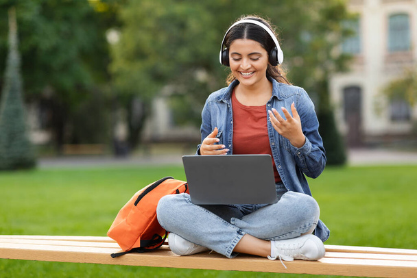 Online education, webinar, course. Happy beautiful young indian woman student have online class while chilling at park, using laptop and wireless headphones, pass online exam, video call with tutor - Photo, Image