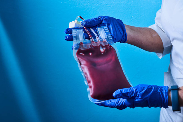 Επιστήμονας φορούν μπλε γάντια χέρι κρατώντας κόκκινη τσάντα αίματος από δότη σε τράπεζα αίματος.Γιατρός και φρέσκο αίμα για τη μετάγγιση.. - Φωτογραφία, εικόνα