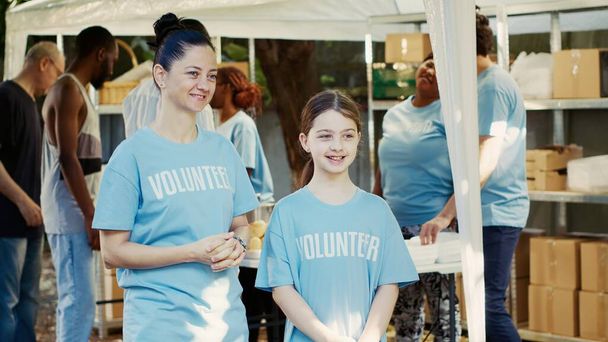Side-view portret van een moeder en dochter ter ondersteuning van een voedselbank buiten campagne om honger te bestrijden. Twee blanke vrouwen in blauwe vrijwillige t-shirts zijn bereid de daklozen te helpen.. - Foto, afbeelding