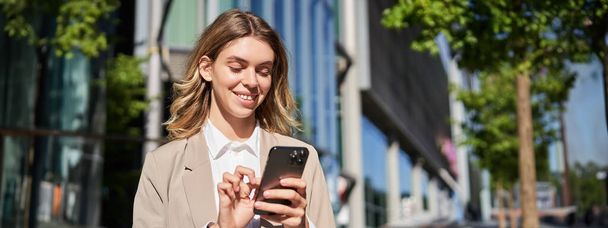Retrato de una hermosa mujer de negocios sonriente usando su teléfono móvil en la calle. Mujer corporativa toque su teléfono inteligente, mensajería, de pie en la calle. - Foto, Imagen