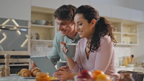Pareja romántica de compras en línea en el desayuno familiar de cerca. Felices cónyuges sonrientes tomando café con croissant mirando en la pantalla de la tableta de la cocina moderna. Par alegre usando gadget digital - Imágenes, Vídeo