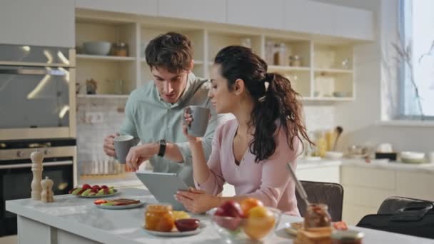 Mari embrassant femme au revoir aller au travail. Joyeux époux souriants buvant le café du matin ensemble sur la cuisine moderne avant le travail. Détendu belle femme tenant tablette ordinateur assis comptoir. - Séquence, vidéo