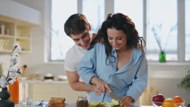 愛情あふれるカップルがキッチンで料理をし,一緒に笑い合った. 若い配偶者を愛し,朝の健康的な朝食を準備する楽しさを持っています. 笑顔の妻は夫の助けを借りて新鮮なリンゴをスライス.  - 映像、動画