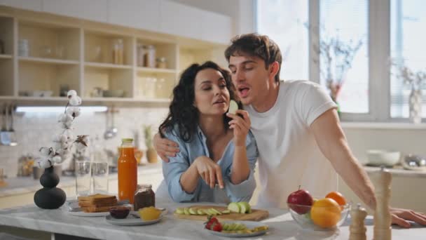 Usmívající se přítelkyně krmení přítele s jablkem plátek v bytě kuchyně zblízka. Smějící se romantický pár baví spolu jíst čerstvé ovoce. Šťastní manželé si užívají rodinného rána doma. - Záběry, video