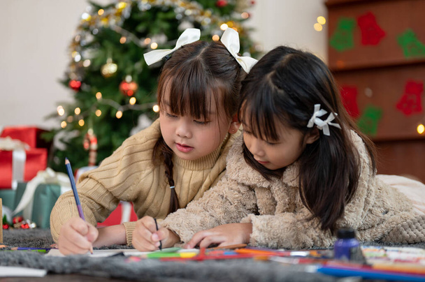 Due adorabili e felici ragazze asiatiche si stanno divertendo a colorare i loro biglietti di Natale con colori a matita, divertendosi e festeggiando il Natale a casa insieme.. - Foto, immagini