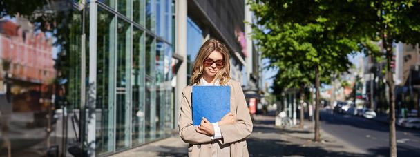 Güneş gözlüklü ve bej takım elbiseli kurumsal bir kadının portresi, elinde ofis belgeleriyle mavi dosya, işe gitmek, güneşli bir günde sokakta yürümek.. - Fotoğraf, Görsel