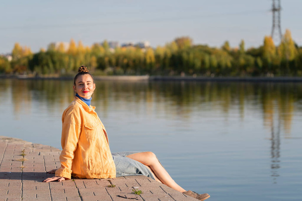 Una joven se sienta junto al río en el terraplén. Concepto: libertad, disfrutar de la vida, estar a solas contigo mismo. Foto de alta calidad - Foto, imagen
