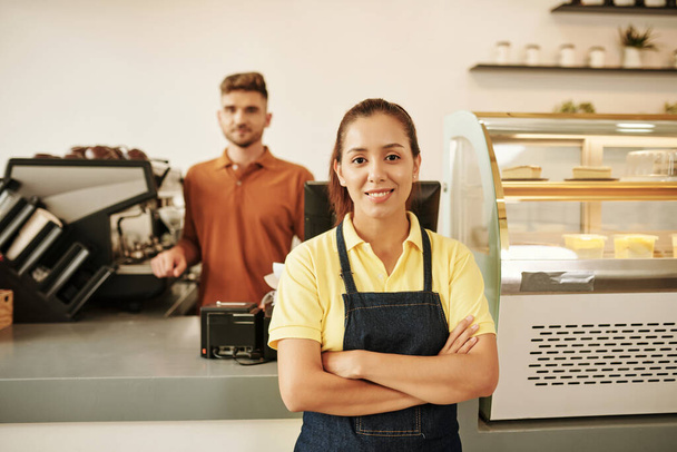 Portret uśmiechniętej kelnerki stojącej przy ladzie z skrzyżowanymi ramionami - Zdjęcie, obraz