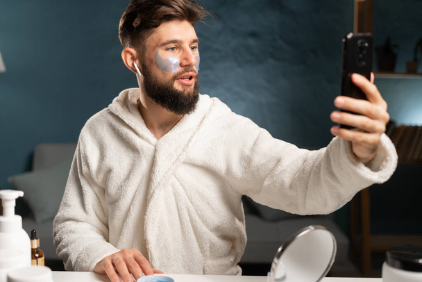 Bearded millennial man dressed in bathrobe records video blog tells about cosmetic products in home interior. Blogging, skincare and cosmetology concept - Photo, Image