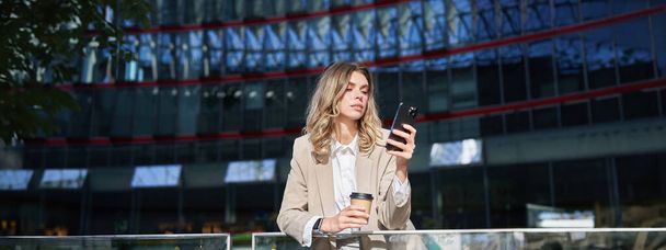 Zakenvrouw in pak met smartphone op straat, koffie drinken, mobiele telefoon app, surfen op internet, een dagje uit. - Foto, afbeelding