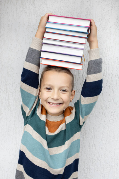 Veselý osmiletý chlapec drží na hlavě hromádku knih. Zpátky do školy. Den knih. Čti s radostí. Vzdělávání, rozvoj představivosti - Fotografie, Obrázek