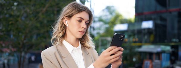 Mujer de negocios exitosa en traje beige, auriculares inalámbricos, mirando el teléfono móvil, utilizando la aplicación para teléfonos inteligentes, de pie al aire libre en la calle. - Foto, Imagen