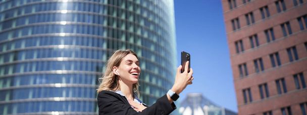 Πορτρέτο της χαμογελαστή εταιρική γυναίκα βιντεοκλήση στο δρόμο, κρατώντας το κινητό τηλέφωνο και χαιρετώντας κάμερα smartphone, φορώντας κοστούμι. - Φωτογραφία, εικόνα