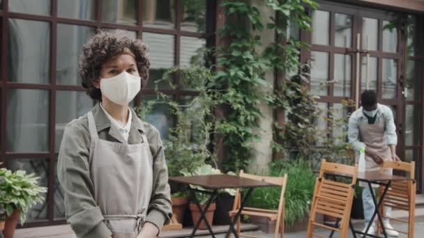 Medium portrait of female Caucasian cafe worker wearing mask standing outside and African American coworker arranging outdoor seatings at background - Footage, Video