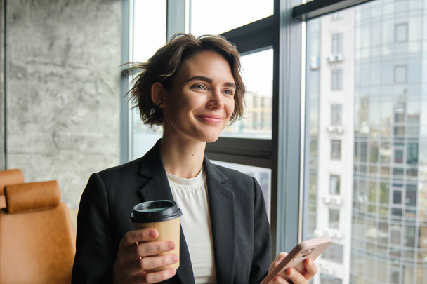 Πορτρέτο της εταιρικής γυναίκα ανάγνωση μήνυμα στο κινητό τηλέφωνο, πίνοντας καφέ, στέκεται κοντά στον καθρέφτη του γραφείου και χαμογελώντας. - Φωτογραφία, εικόνα