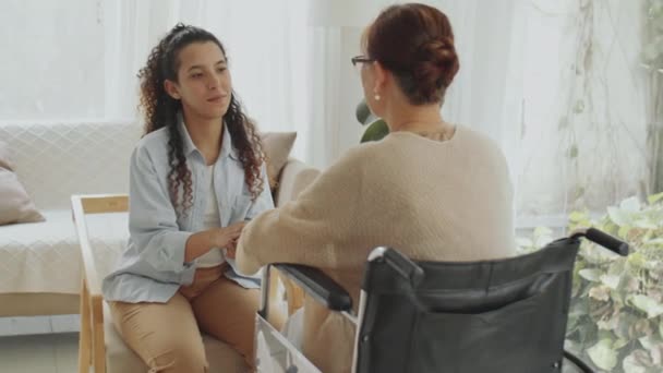 Νεαρή γυναίκα φροντίστρια κρατιέται χέρι-χέρι με ηλικιωμένη γυναίκα σε αναπηρική καρέκλα και της μιλάει στο σαλόνι ενώ την υποστηρίζει κατά τη διάρκεια της ημέρας - Πλάνα, βίντεο