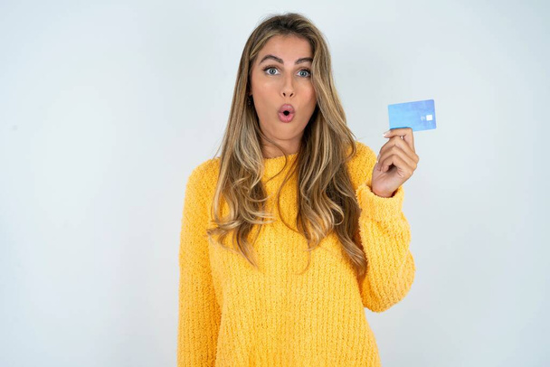 Foto de una joven mujer caucásica con suéter amarillo asombrada y sorprendida sosteniendo tarjeta de crédito - Foto, imagen