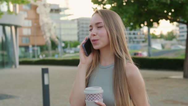 Mujer joven feliz con el pelo largo y suelto rubia pasa tiempo en el parque hablando con el novio por teléfono. Lady se ríe alegremente bebiendo café al aire libre - Metraje, vídeo