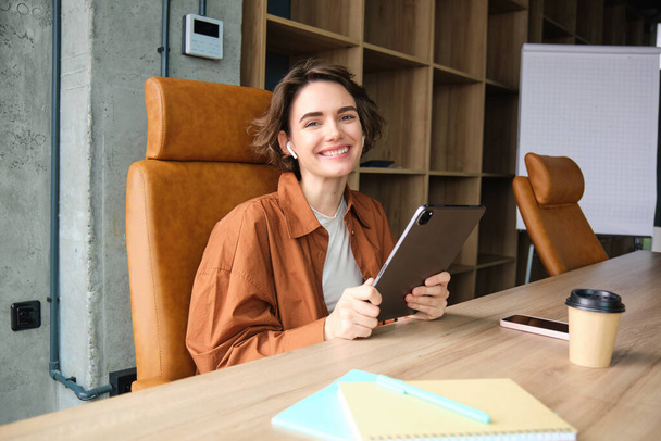Retrato de la mujer trabajadora en la oficina, sentada en la mesa con la tableta digital, usando auriculares inalámbricos, sonriendo a la cámara. - Foto, Imagen