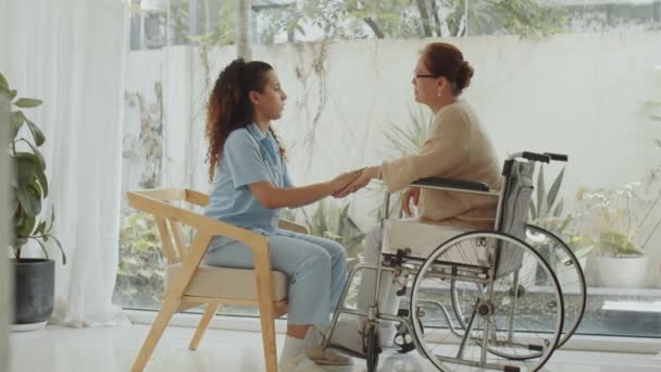 Full shot of female doctor in medical uniform holding hands and talking with senior Asian woman in wheelchair while visiting her at home - Footage, Video