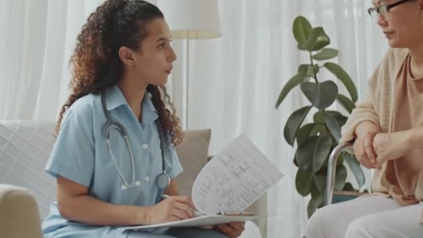 Colpo inclinato di giovane medico donna in uniforme medica che prende appunti e discute con la paziente anziana sulla sedia a rotelle durante la consultazione medica a casa - Filmati, video