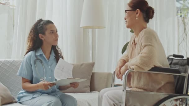 Mediana toma completa de mujer asiática mayor en silla de ruedas explicando los síntomas a la doctora durante la consulta de salud en casa - Imágenes, Vídeo