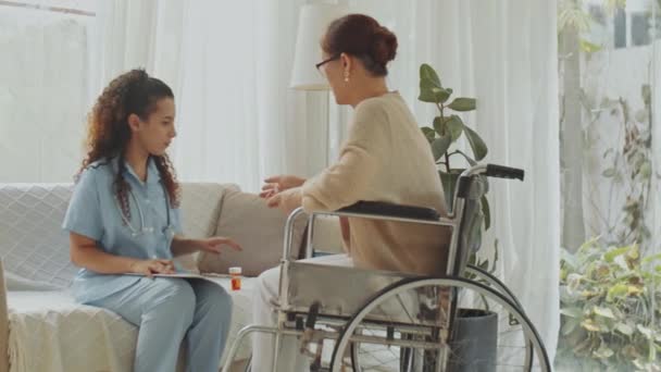 Молода жінка-лікар розповідає про ліки старшій жінці в інвалідному візку і пояснює план лікування під час домашньої консультації - Кадри, відео