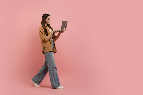 Счастливая девочка-подросток с макушкой в руках, шагающая по розовому студийному фону, полная длина смокинга, молодая женщина, использующая его для интернет-покупок или дистанционного копирования, пространство для копирования - Фото, изображение