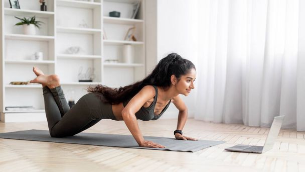 Sport, Fitness, gesunder Lebensstil. Motiviert sportlich attraktive schlanke junge Frau in grüner Sportbekleidung, die zu Hause auf der Yogamatte trainiert, Blick auf Laptop-Bildschirm, Panorama mit Kopierraum - Foto, Bild