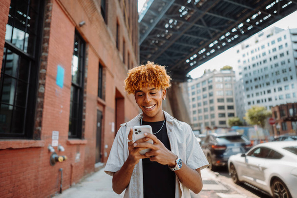 Yakışıklı Afro-Amerikan bir gencin portresi biseksüel şehirde caddede yürüyor, akıllı bir telefona mesajlar yazıyor, gülümsüyor ve poz veriyor. - Fotoğraf, Görsel