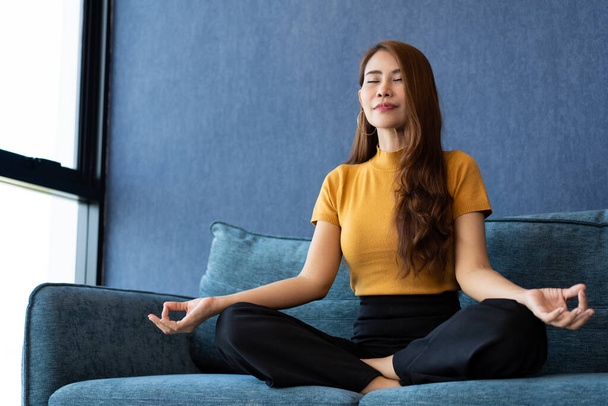 Ritratto di donna asiatica sana che medita ed esercita la posa yoga sul divano al mattino. Rilassamento zen e stile di vita consapevole, concetto di allenamento indoor per una buona pratica sana e respiratoria. - Foto, immagini